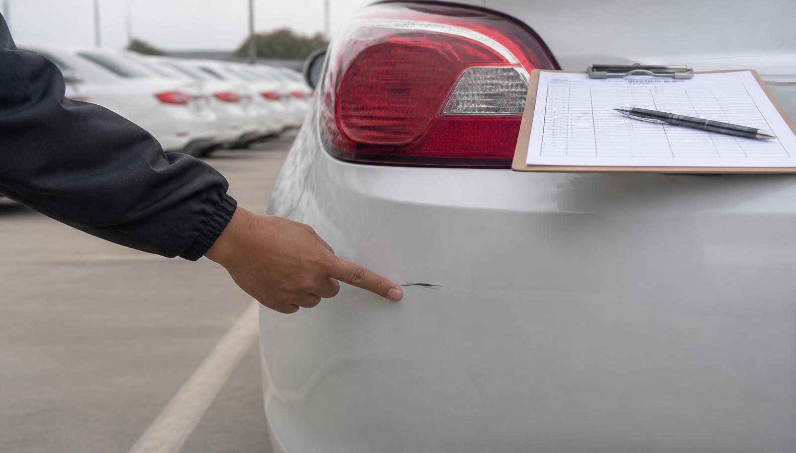 A hand pointing to a small scratch on the rear bumper of a white sedan in a rental parking lot, beside a blank inspection checklist on a clipboard.